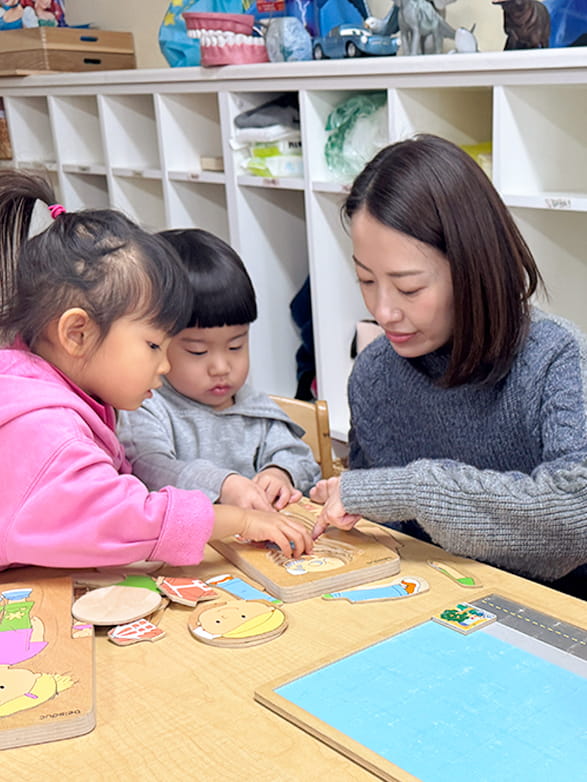 外国人のイラストが描いているパズルを子どもたちを補助しながら一緒に作っていく保育士の女性