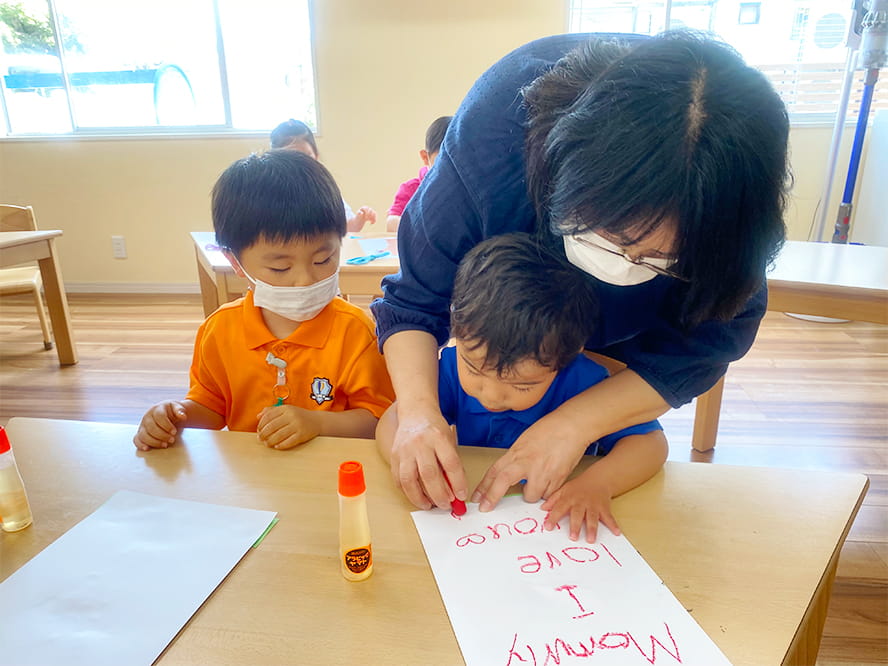 カラフルなクレヨンを使い、英会話の単語を紙に書いている生徒さんと優しく見守る女性保育士