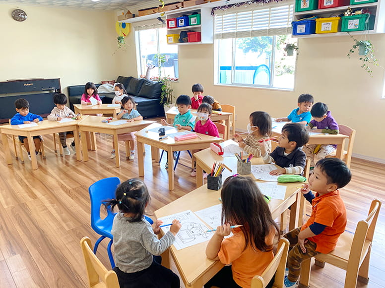 一つずつ椅子に座り、紙とペンを持って塗り絵をしているキンダーガーテンの生徒たち