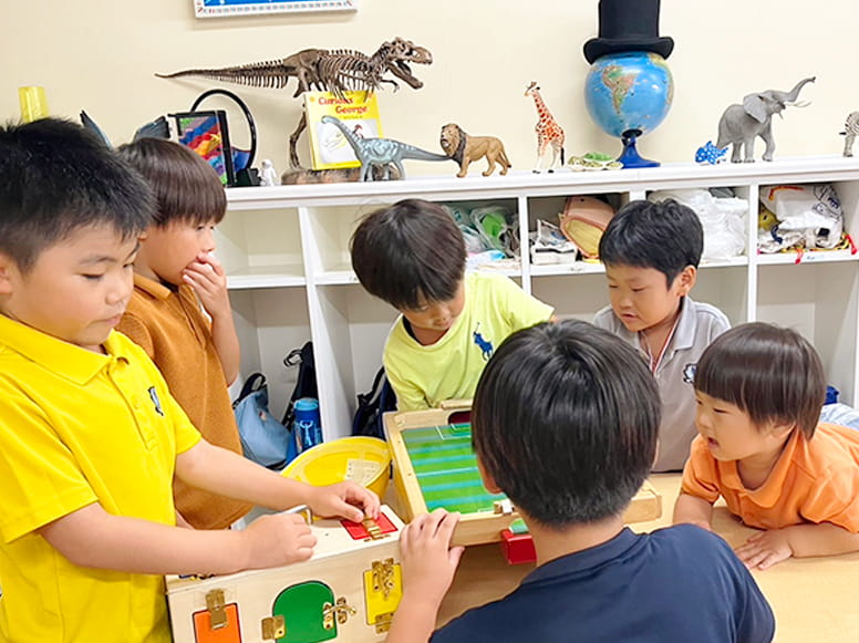 ロッカーの上に恐竜などの模型が置かれた教室で木のおもちゃなどを使い話をしながら遊ぶ様子